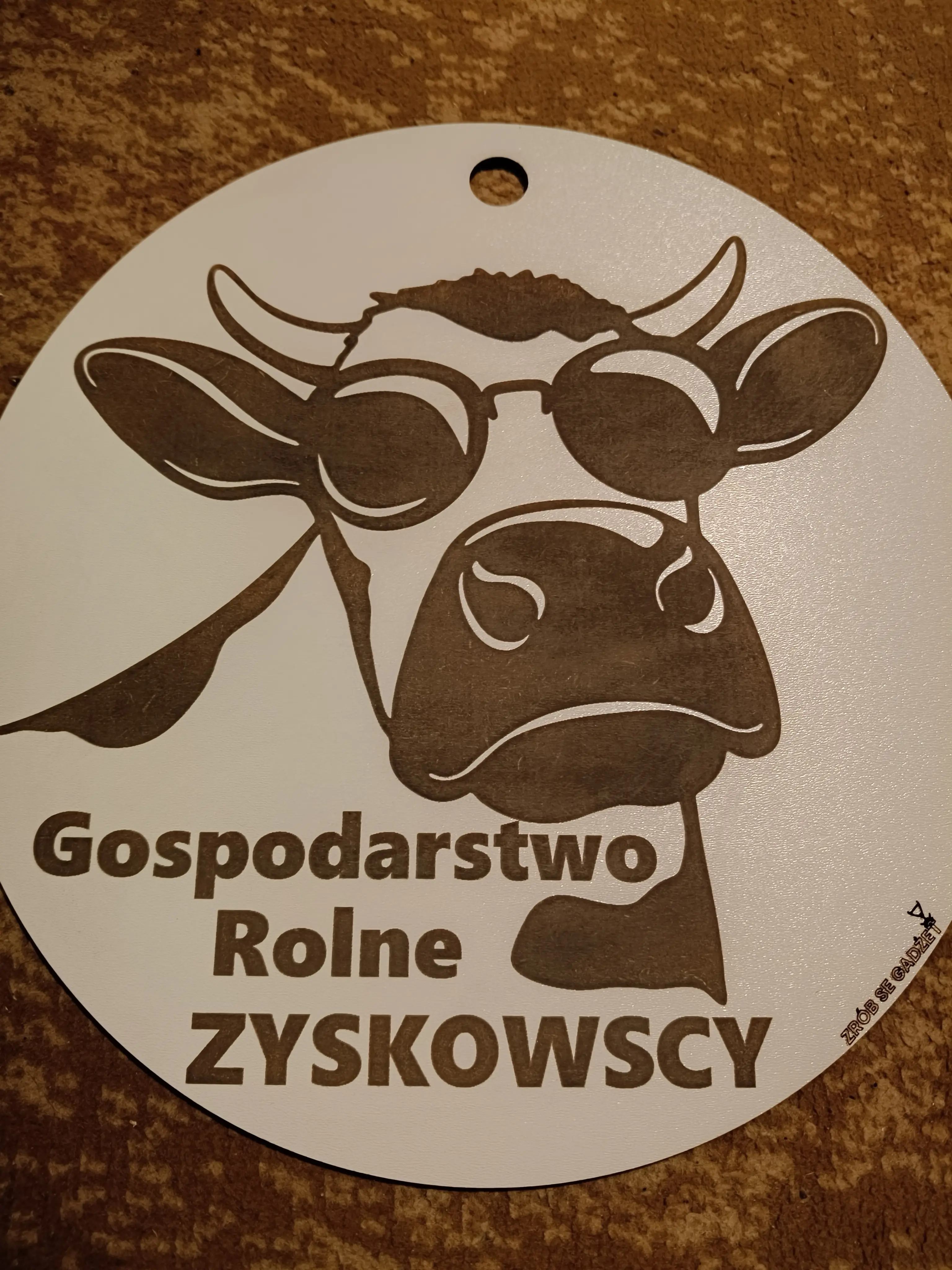 Okrągły szyld z ilustracją krowy w okularach przeciwsłonecznych i napisem Gospodarstwo Rolne ZYSKOWSCY